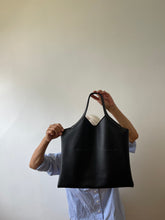 Load image into Gallery viewer, a person is standing holding a black leather bag with short handle and a short v shaped dip at the center of the bag. the black leather used looks supple.
