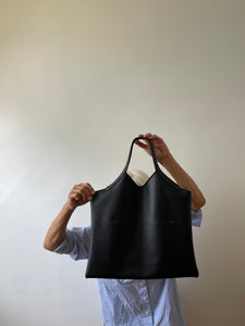 a person is standing holding a black leather bag with short handle and a short v shaped dip at the center of the bag. the black leather used looks supple.
