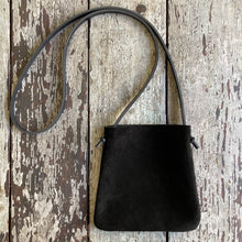 Load image into Gallery viewer, standing x-small black suede bag lays flat on a tabletop. its bottom corners are rounded. strap length can be worn crossbody. This bag is artisan made and handcrafted.
