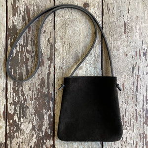 standing x-small black suede bag lays flat on a tabletop. its bottom corners are rounded. strap length can be worn crossbody. This bag is artisan made and handcrafted.