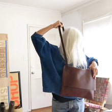 Load image into Gallery viewer, Woman stands moving the leather strap of a brown leather bag over her head. The medium sized bag has a front flap that stops midway. 
