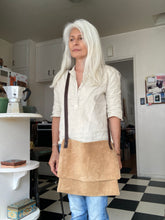 Load image into Gallery viewer, Woman standing wears a medium-size tan suede crossbody bag with dark brown leather straps. it’s flap is closed. The suede looks supple.
