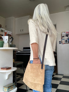 Woman standing wears a medium-size tan suede crossbody bag with dark brown leather straps. it’s flap is closed. The suede looks supple.