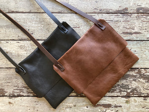 Two leather bags of the same design lay flat on a table top— one is black, the other a distressed brown. Both of their flaps are closed. The shoulder straps are tied and knotted to the top of the bag.