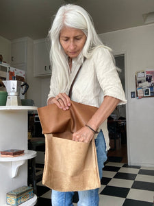Woman standing wears a medium-size tan suede crossbody bag with a dark brown leather strap. She holds the bags flap open reaching inside for something.