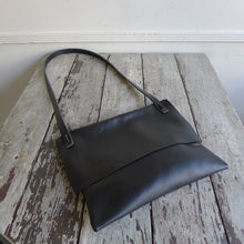 Load image into Gallery viewer, A black leather rectangular shaped bag lays flat on a table top. Its flap is closed. The shoulder strap is tied and knotted to the top of the bag.
