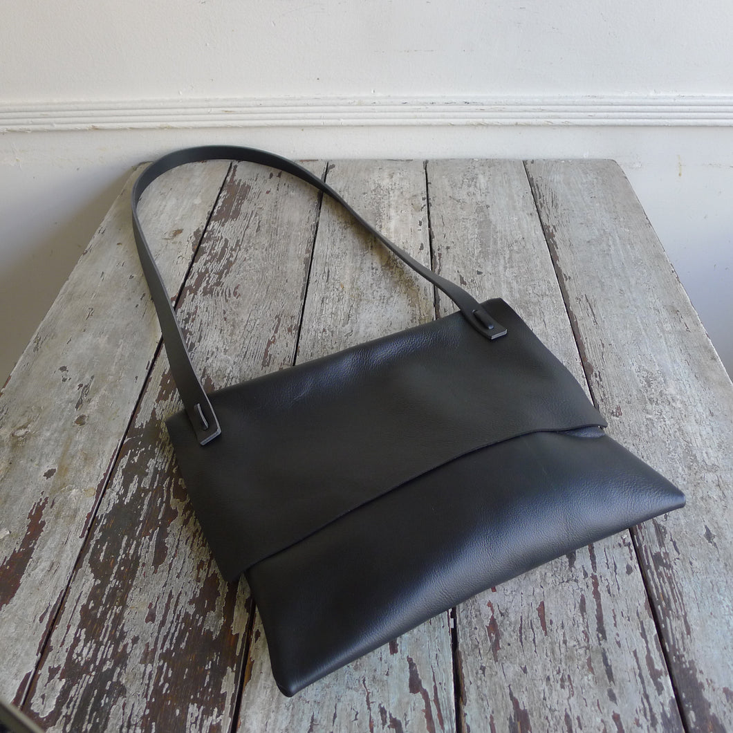 A black leather rectangular shaped bag lays flat on a table top. Its flap is closed. The shoulder strap is tied and knotted to the top of the bag.