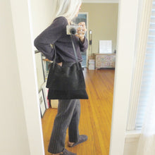 Load image into Gallery viewer, Woman standing. She wears a medium-size black suede bag over her shoulder. it’s flap is closed. 
