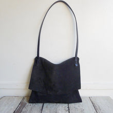 Load image into Gallery viewer, a medium-size black suede bag with its flap closed sits on a table top. Its crossbody length leather strap is fully extended.
