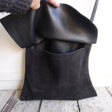 Load image into Gallery viewer, A medium-size black suede bag sits on a table top. A hand holds its flap up showing the inside of the bag. The combination of suede &amp; leather looks very soft and supple.
