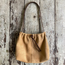 Load image into Gallery viewer, A photograph of a small tan suede bag fully gathered at the top tied with a leather tie knotted for closure . It’s short brown strap is shoulder length. The bag has a center seam and gusset bottom.
