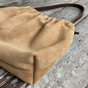 Close up of a small tan suede bag’s gusseted bottom. The bag’s top is fully gathered and has a short brown shoulder length strap.