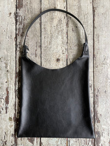 An all-black shoulder length leather bag lays flat on a table top. The top of the bag dips down into a half-circle shape. Leather straps are tied and knotted at the top left and right sides. The bottom half of the bag is a rectangular shape and the leather body looks supple.