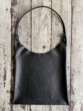 Load image into Gallery viewer, An all-black shoulder length leather bag lays flat on a table top. The top of the bag dips down into a half-circle shape. Leather straps are tied and knotted at the top left and right sides. The bottom half of the bag is a rectangular shape and the leather body looks supple.
