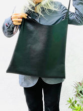 Load image into Gallery viewer, Woman showing an all-black leather bag. The top of the bag dips down into a half-circle shape. Leather straps are tied and knotted at the top left and right sides. The bottom half of the bag is a rectangular shape and the body is flat. The leather looks supple.
