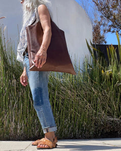 Woman wearing a large size brown leather bag with a short handle around her shoulder. The leather looks soft.