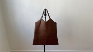 Pictured hanging from a stand is a distressed brown leather bag with short handle and a short v shaped dip at the center of the bag. the leather looks supple.