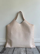 Load image into Gallery viewer, sitting on a table is a bag with short handles and a short v shaped dip at the center of the bag made in a natural veg tan leather. no patina yet. 
