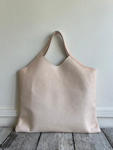 sitting on a table is a bag with short handles and a short v shaped dip at the center of the bag made in a natural veg tan leather. no patina yet. 