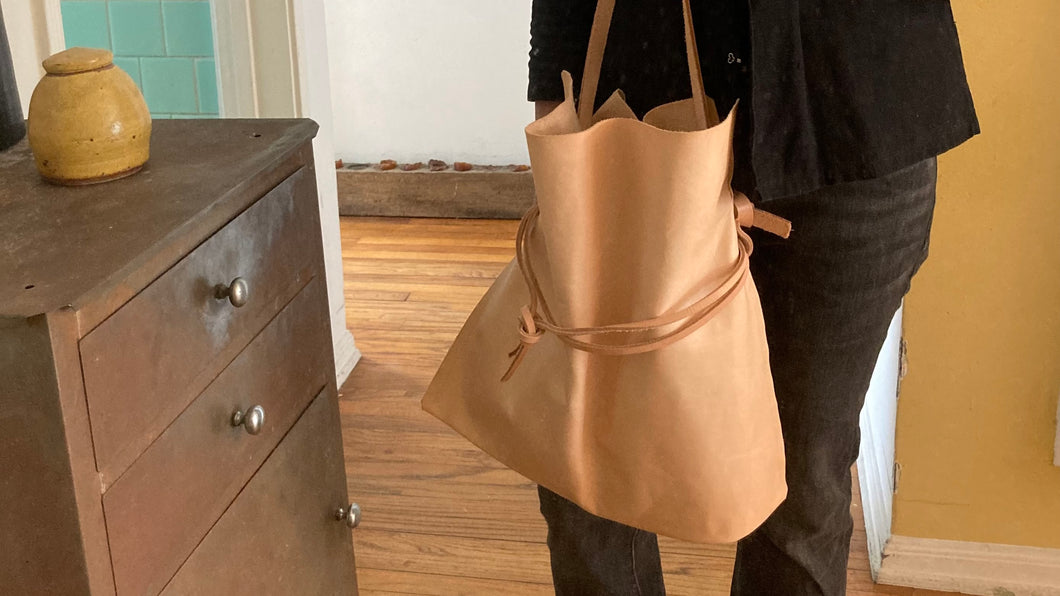 Close up of natural vegetable tanned leather bag (with a light patina). Drawstring style— veg tan leather ties that cinch in for closure or remove ties to wear without. Made with knotted/adjustable crossbody length straps.