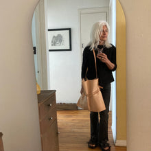 Load image into Gallery viewer, Woman standing wearing a natural vegetable tanned leather bag (with a light patina) over her shoulder. Drawstring style— veg tan leather ties cinch in for closure. Ties are removable to wear bag to its fully extended width. The crossbody length the strap is adjustable.
