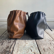 Load image into Gallery viewer, Close up of a side view of two small leather bags of exact style sitting side by side— one in tan and the other in black. Each bag is fully gathered at the top tied with leather ties knotted for closure. Each bottom is gusseted.
