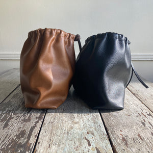 Close up of a side view of two small leather bags of exact style sitting side by side— one in tan and the other in black. Each bag is fully gathered at the top tied with leather ties knotted for closure. Each bottom is gusseted.