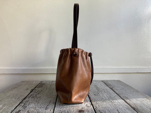 Close up of a side view of a small tan leather bag fully gathered at the top tied with a leather tie knotted for closure. its brown shoulder strap tied and knotted to both sides. The bag’s bottom is gusseted.