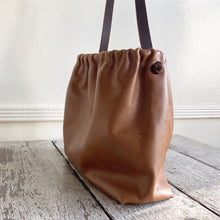 Load image into Gallery viewer, Close up of a side view of a small tan leather bag fully gathered at the top tied with a leather tie knotted for closure. its brown shoulder strap tied and knotted to both sides. The bag’s bottom is gusseted.
