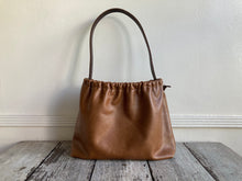 Load image into Gallery viewer, A photograph of a small tan leather bag fully gathered at the top. It’s short brown strap is shoulder length and tied to each side. The bag’s bottom is gusseted.
