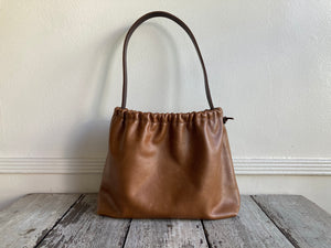 A photograph of a small tan leather bag fully gathered at the top. It’s short brown strap is shoulder length and tied to each side. The bag’s bottom is gusseted.