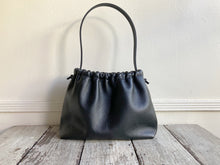 Load image into Gallery viewer, A small black leather bag fully gathered at the top tied with a leather tie knotted for closure. It’s short black leather strap is shoulder length. The bag’s bottom is gusseted.
