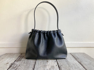 A small black leather bag fully gathered at the top tied with a leather tie knotted for closure. It’s short black leather strap is shoulder length. The bag’s bottom is gusseted.