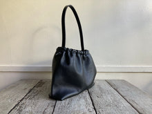 Load image into Gallery viewer, Close up of a side view of a small black leather bag fully gathered at the top. its black shoulder strap tied and knotted to both sides. The bag’s bottom is gusseted.
