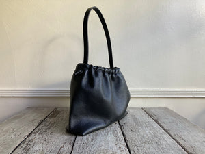 Close up of a side view of a small black leather bag fully gathered at the top. its black shoulder strap tied and knotted to both sides. The bag’s bottom is gusseted.
