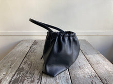 Load image into Gallery viewer, Close up of a side view of a small black leather bag fully gathered at the top. its black shoulder strap tied and knotted to both sides. The bag’s bottom is gusseted.
