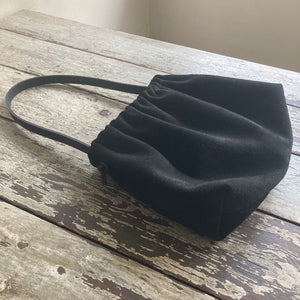 A photograph of a small black suede bag fully gathered at the top. It’s short black leather strap is shoulder length and tied to each side. The bag’s bottom is gusseted.