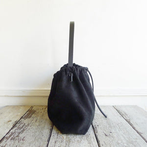 Close up of a small black suede bag fully gathered at the top and tied on one side. its black shoulder strap tied and knotted to both sides. The bag’s bottom is gusseted.