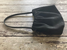 Load image into Gallery viewer, A photograph of a small black suede bag fully gathered at the top. It’s short black leather strap is shoulder length. The bag’s bottom is gusseted.
