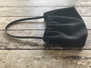 A photograph of a small black suede bag fully gathered at the top. It’s short black leather strap is shoulder length. The bag’s bottom is gusseted.