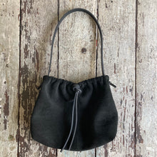 Load image into Gallery viewer, A photograph of a small black suede bag fully gathered at the top tied with a leather tie knotted for closure. It’s short black leather strap is shoulder length. The bag has a center seam and a gusset bottom.
