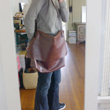 Load image into Gallery viewer, Person standing wearing a brown leather crossbody messenger-style bag over her shoulder. The large-size bag has a long flap and a gusset bottom. The leather appears rich in color and supple.
