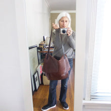 Load image into Gallery viewer, whited haired woman holding by its strap a brown leather crossbody messenger-style bag over her shoulder. The large-size bag has a long flap and a gusset bottom. The leather appears rich in color and supple.
