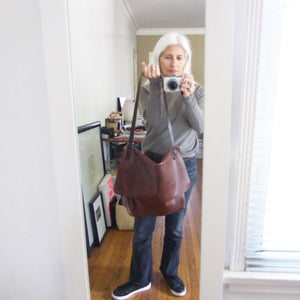 whited haired woman holding by its strap a brown leather crossbody messenger-style bag over her shoulder. The large-size bag has a long flap and a gusset bottom. The leather appears rich in color and supple.