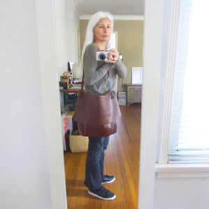 whited haired woman wearing a brown leather crossbody messenger-style bag over her shoulder. The large-size bag has a long flap and a gusset bottom. The leather appears rich in color and supple
