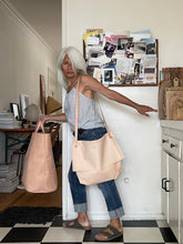 Load image into Gallery viewer, whited haired woman wearing a natural vegetable tanned leather crossbody messenger-style bag over her shoulder appearing to be in a hurry to get out the door. The large-size bag has a long flap and a gusset bottom. a very light patina to the leather.
