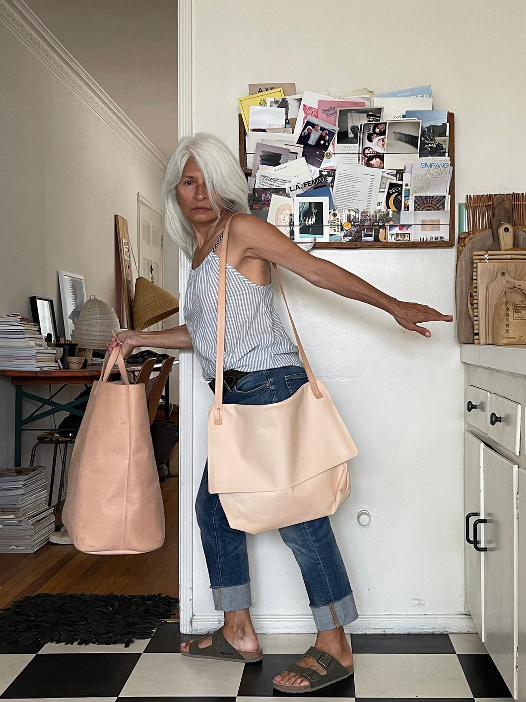 whited haired woman wearing a natural vegetable tanned leather crossbody messenger-style bag over her shoulder appearing to be in a hurry to get out the door. The large-size bag has a long flap and a gusset bottom. a very light patina to the leather.