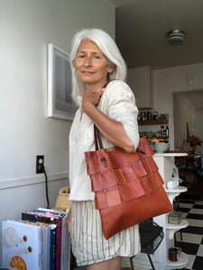 A saffron colored leather bag with three rows of wide color matched leather hand sewn fringe covering 1/3 of the small-size bag starting at the top is worn by a white haired woman looking into the camera. Its shoulder length straps are dark brown in color.
