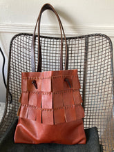 Load image into Gallery viewer, A saffron colored leather bag with three rows of matching wide leather hand sewn fringe covering 1/3 of the small-size bag sits on a chair. Its shoulder length leather straps are a dark brown color.
