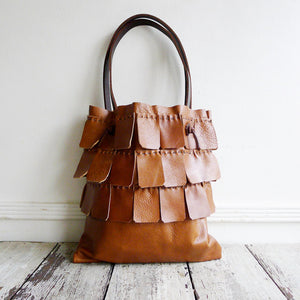 A saffron colored leather bag with three rows of matching wide leather hand sewn fringe covering 1/3 of the small-size bag sits on a table top. Its shoulder length leather straps are a dark brown color.